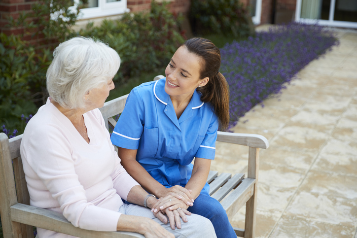 Senior Woman Sitting On Bench And Talking With Nurse In Retirement Home